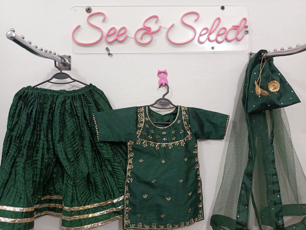 Green traditional outfit with gold embroidery on a white wall with 'See & Select' sign.